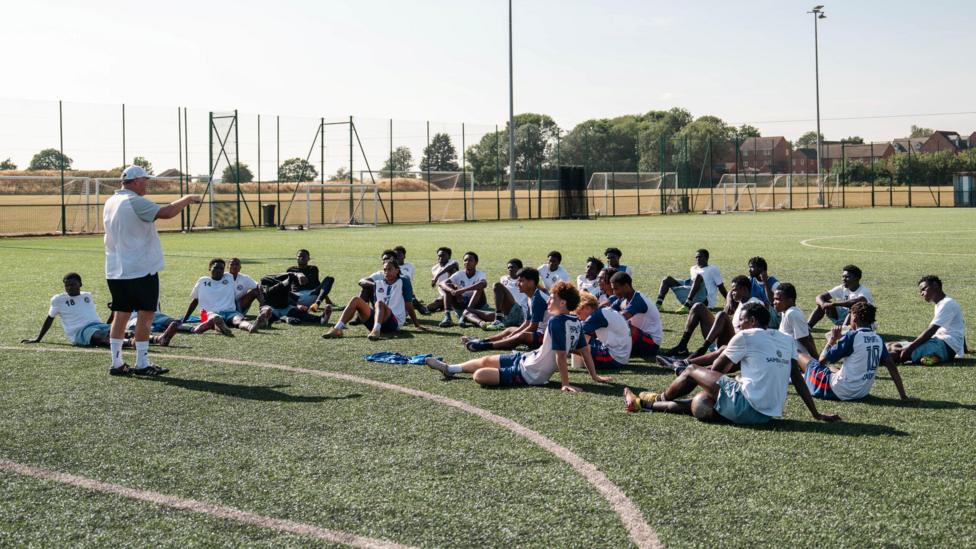 Coach Rob Newman stands on an artificial football pitch and points while giving instruction to around 30 players from the Samba Stars academy who are sitting on the ground in a group in front of him. A fence runs around the pitch and further training facilities, including white goals, can be seen in the background