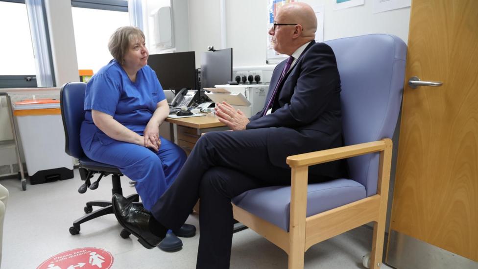 First Minister John Swinney during a visit to officially open the new GP walk-in service at Randolph Wemyss Memorial Hospital in Leven, Fife, as he announces the next locations for the service - A suited visitor and a healthcare worker in blue scrubs sit facing each other in a GP consultation room. The room contains computer monitors, a desk, clinical equipment and a chair, with daylight coming through windows in the background.