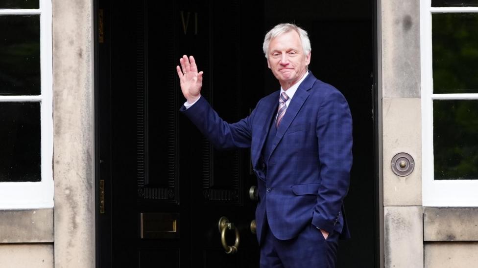 Ivan McKee arrives at Bute House, Edinburgh - A person wearing a blue suit stands in the doorway of a stone-fronted building, raising one hand in a wave. The black entrance door is framed by large windows and stone columns.
