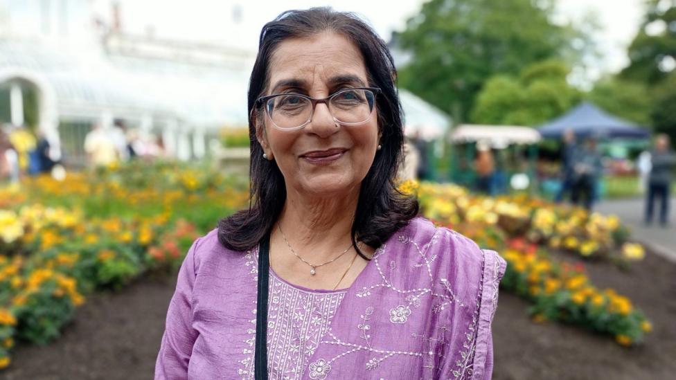 Nisha wears a purple kurta and has black hair. She is wearing glasses and stands in front of a botanical garden.