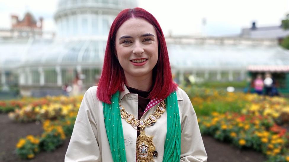 Róis-Máire has hair dyed reddish-purple. Around her neck is a green scarf. Behind her, botanical gardens.