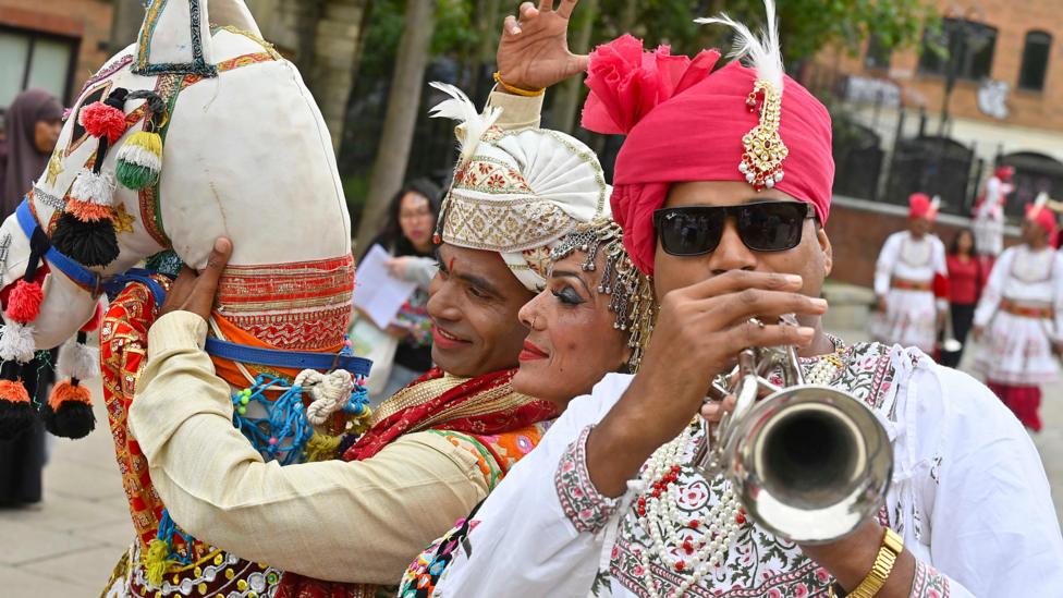A man is playing a silver trumpet. He is wearing a white top with a red turban. Another man holding a plastic horse is on the left side of the image with a woman in the middle.