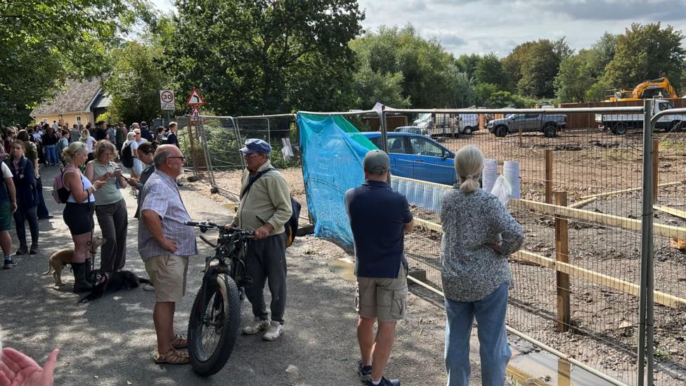 A crowd of people standing behind a metal fence, which is blocking access to the field. Diggers and trucks are at work on the land.