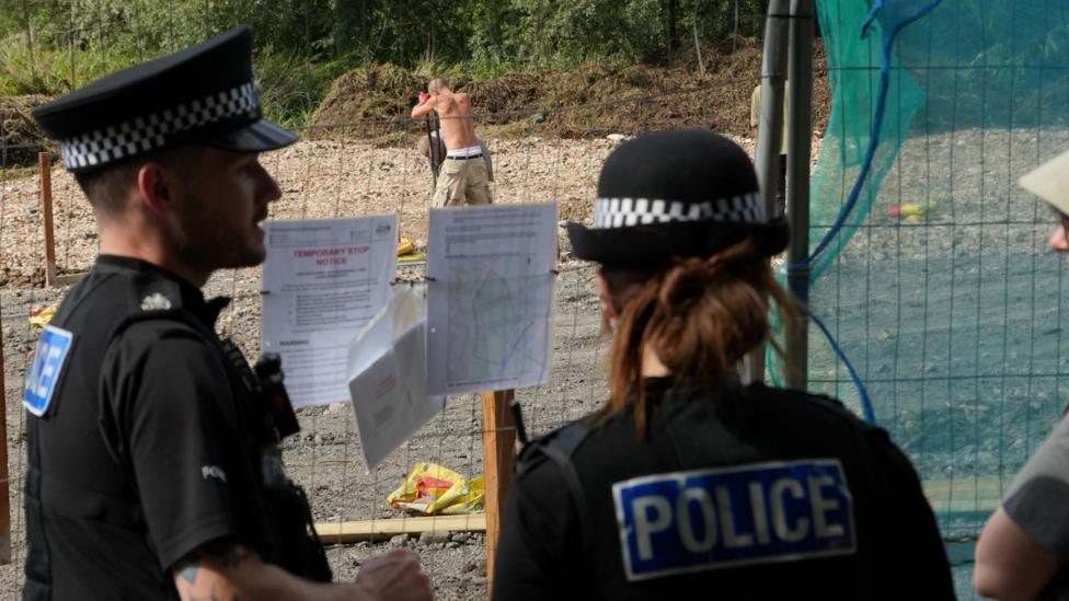Two police officers watching on as a shirtless man digs on the field, which is covered with hardcore.