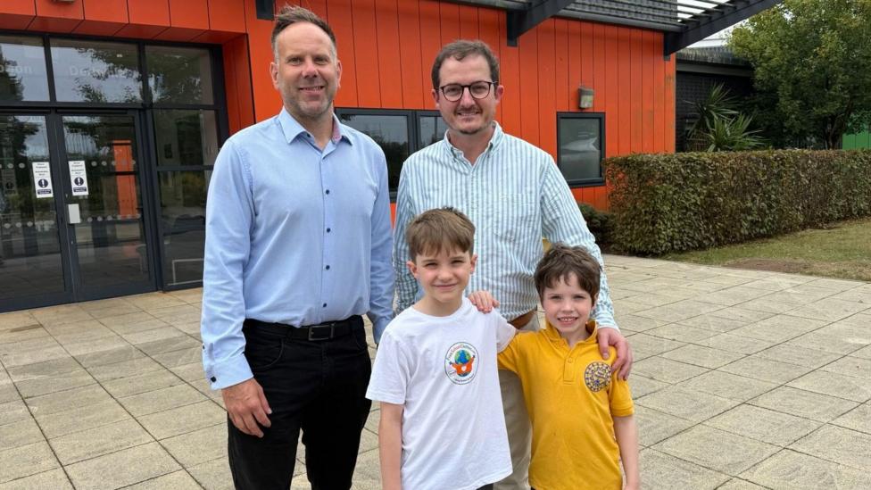 Deputy head teacher, Gareth Evans wears a blue shirt and black trousers and is pictured next to Jon who is wearing a striped shirt and cream trousers and his two children. 10 year old, Tom wears a white T-shirt with Free school clothes logo on it and his 6 year old brother, Sion, wears a school yellow polo shirt