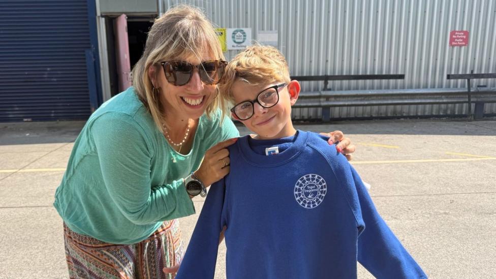 Elise wears a green top, and multi coloured trousers and has blonde hair and wears sunglasses. She is pictures next to her 10 year old son, Charlie, holding a blue jumper next to which she got from the free school clothes scheme.