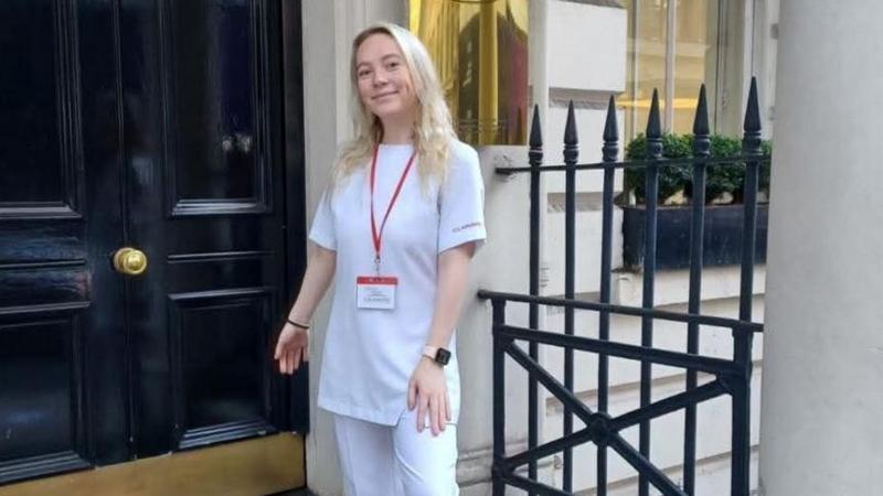 Ffion stood outside a navy door and gate with a gold sign displaying the Clarins logo. She wears a white two-piece uniform with a red lanyard. She has long blonde hair and smiles looking at the camera.