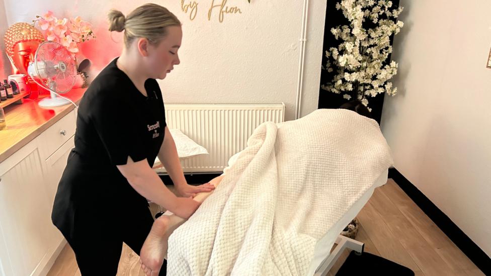 Ffion massaging a female leg on a massage bed in her bright treatment room. A large white blanket is over the woman on the bed making her non identifiable. 