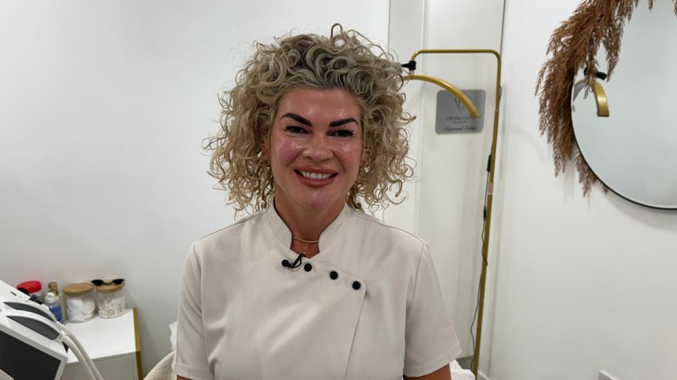 Rhianne sat on a white treatment bed wearing a white spa unifprm. She has wavy blonde hair and smiles looking at the camera. Behind her is a gold mirror, spa mahcinery and white wall. 