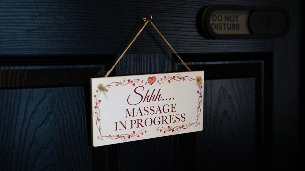 A white sign with a pink floral border which says "Shhh...massage in progress" on a navy door. In the top right corner, there is a do not disturb sign. 