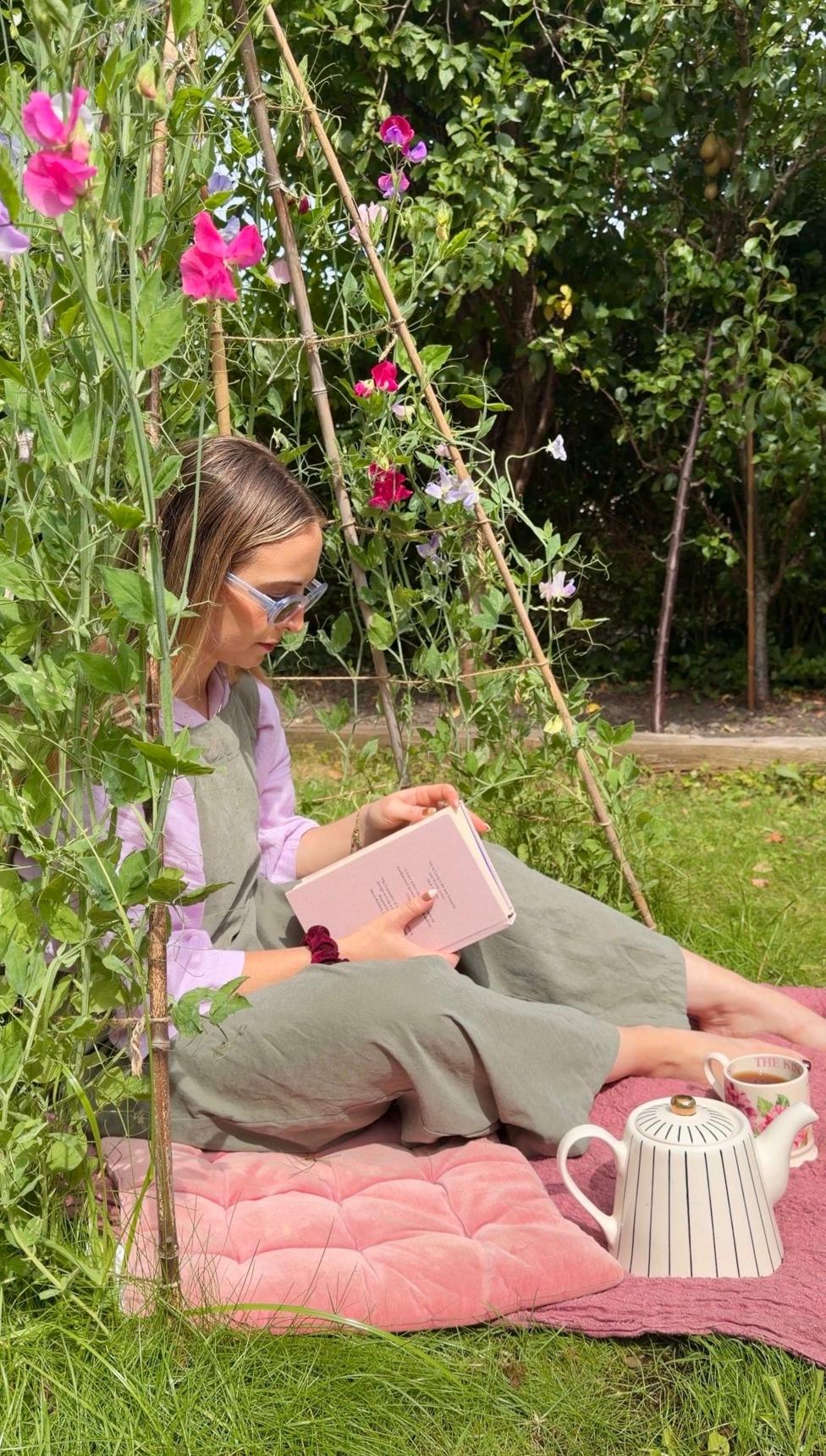 Hannah sitting on a mat in her back garden, reading a book, surrounded by plants