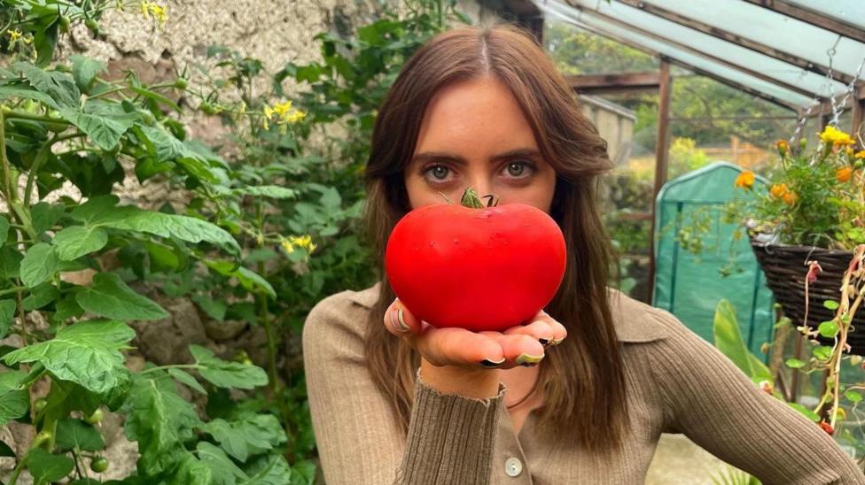 Naomi is pictured her in a greenhouse against a backdrop of flowers. She's holding a large red tomato in front of her face, placed on her palm. She has long brown hair and is wearing a knit green top.
