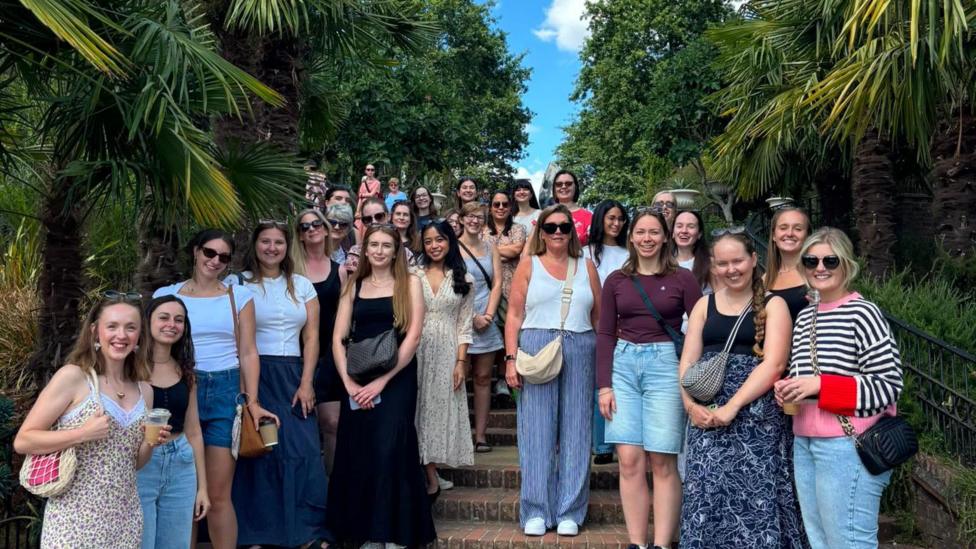 This photograph includes more than 20 people on the steps of a garden. They're all various ages and heights. There are trees lining the steps.