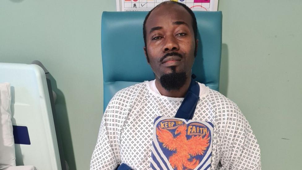 Bernard Agyemang is wearing a patterned hospital gown and is sitting on a chair. He is holding a painted sign which reads: Keep the faith. 