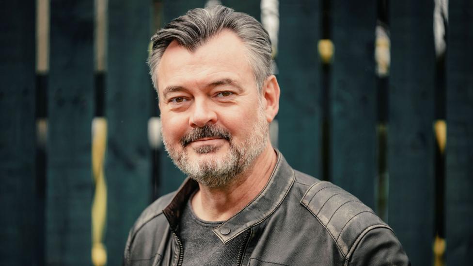 Grant Stott, with a grey beard and wearing a leather jacket, standing against a garden fence