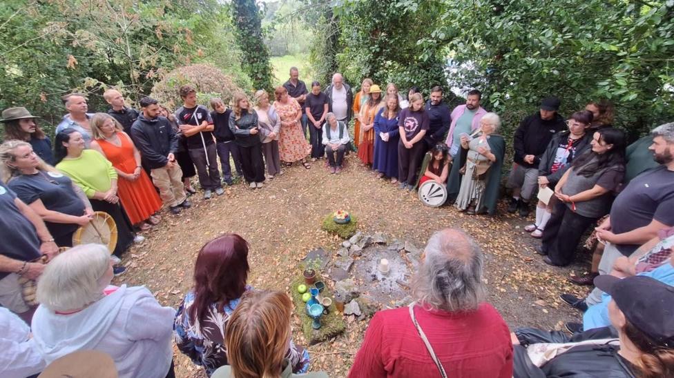 A group of people standing in a circle outdoors. On the ground in front of them are several flat stones. Cups of wine and apples are placed on the stones.