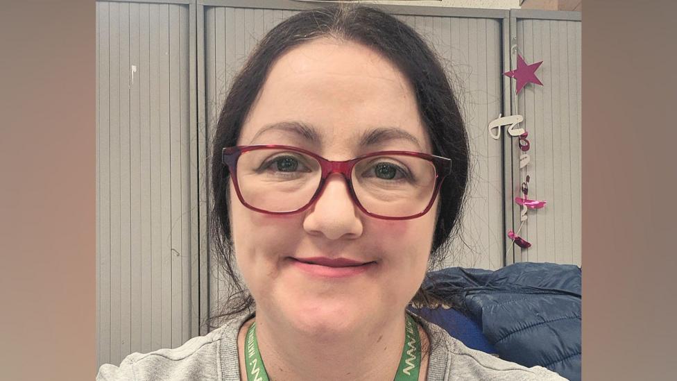Fiona Byrne has dark brown hair pulled back into a low ponytail. She is wearing dark pink glasses and a grey jumper.