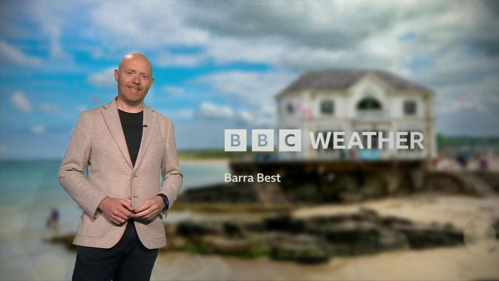 Barra Best is standing in front of a TV studio green screen. Behind him is a blurry picture of a house on a beachfront. It says BBC weather on the screen in white letters.
