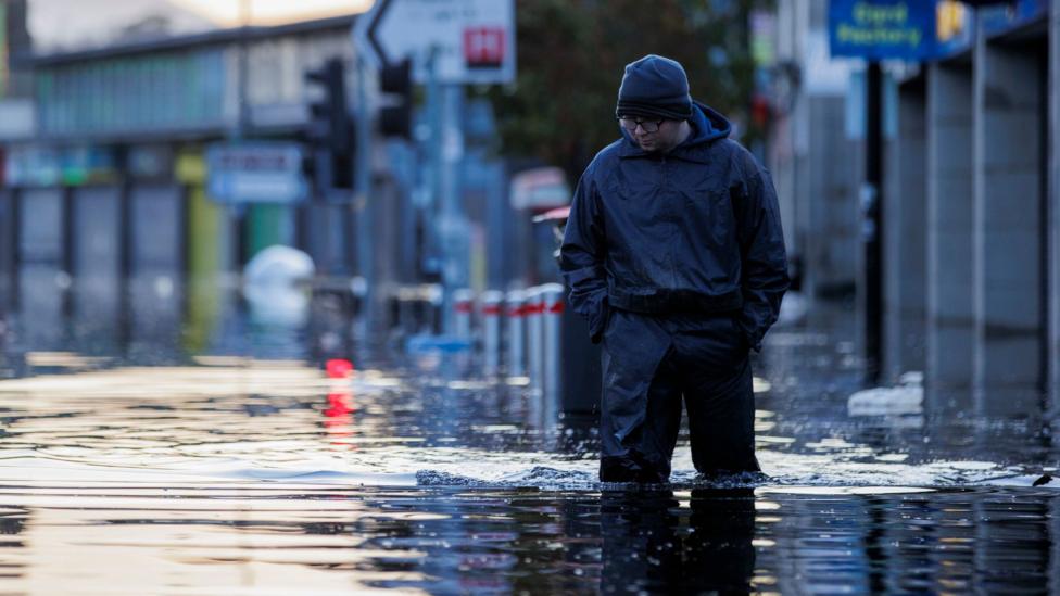 A man wearing dark waterproof overalls and a dark beanie hat wades through knee-high water on Downpatrick's Market Street.