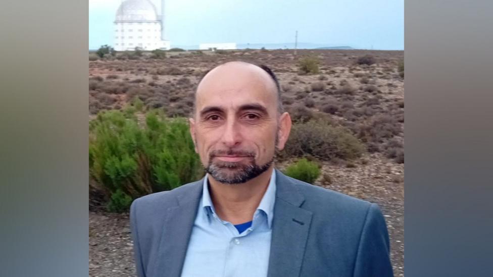 Marc Sarzi is a middle aged man. He's wearing a blue shirt and is standing on scrubland outside a large domed building which contains a telescope.