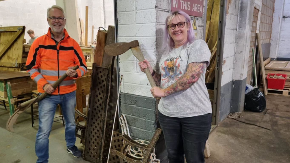 Jim and a woman stand inside their building both holding old rusty axes and laughing. They're leaning by a wall with wood, materials and other items around and behind them.