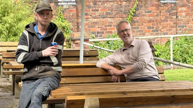Jim and a volunteer sit on benches outside under umbrellas. Jim has a brown jumper on and glasses with a grey beard and the second man has a jumper on with navy trousers. He is sat holding a drink can.