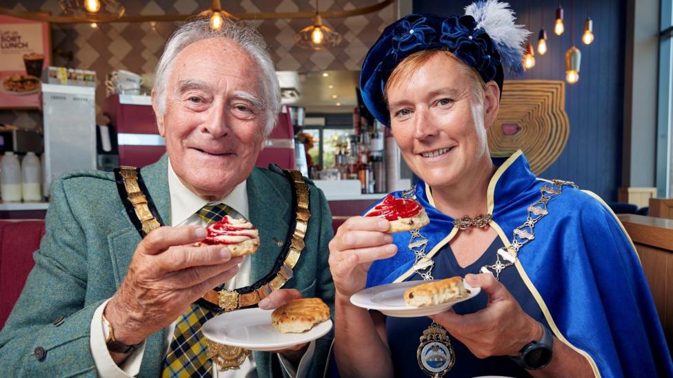 Mayor of Saltash Brian Stoyel and the Mayor of Kingsbridge Julia Wingate try a Costa Coffee 50/50 scone, which has jam and cream piped onto it. Both people are wearing their ceremonial mayoral chains and clothing.