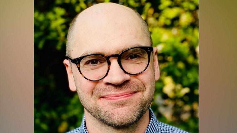 A man called Neil Buttery. He is balding and has a trimmed stubbly beard. He has glasses on. Buttery is stood in front of a hedge.