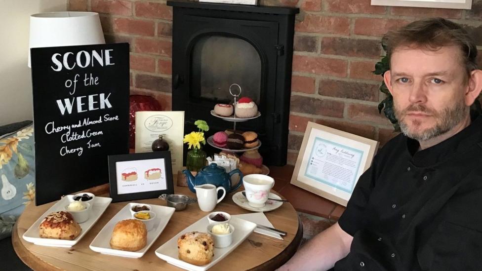 A man called Simon Searle sat next to a table with scones and cream teas on them. There is a blackboard sign which says "scone of the week" on it.