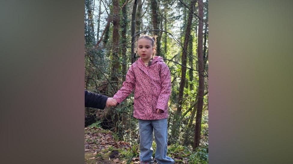 A girl wearing a pink coat looking at the camera. She is standing in a forest.