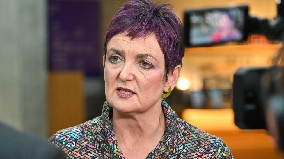 Angela Constance, who has short purple hair, speaks to a journalist in the Scottish Parliament. She is wearing a multi-coloured jacket, with the picture taken over the shoulder of the journalist and from behind a camera.