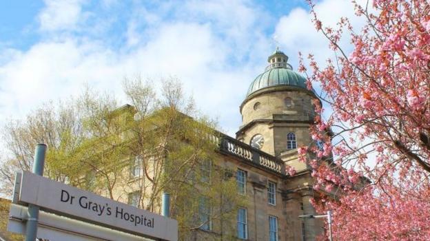 Hospital building with a 'Dr Gray's' sign and some pink blooms on a tree to the right of the image.