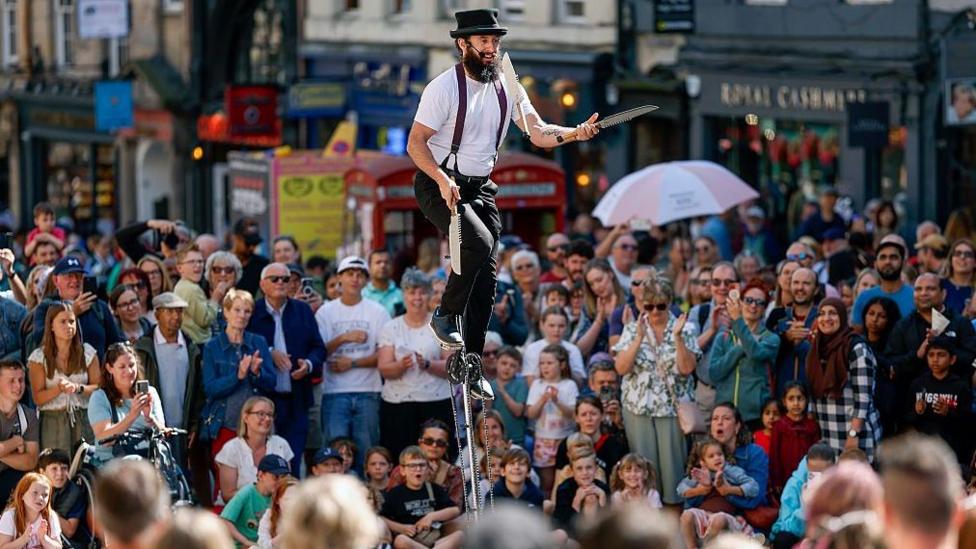 Man on a high unicycle with long knives in his hands, about to thrown them in the air. There is a large crowd watching him on Edinburgh's Royal Mile. He has a beard and is wearing a white T-shirt, black trousers, braces and a black hat.