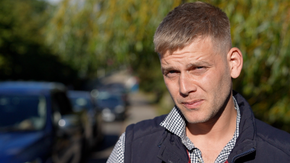 Richard Lines has short light brown hair, and slight stubble. He looks to the camera, standing outside with the green leaves of trees behind him (out of focus), while he wears a navy blue Gillet and black and white checked shirt.