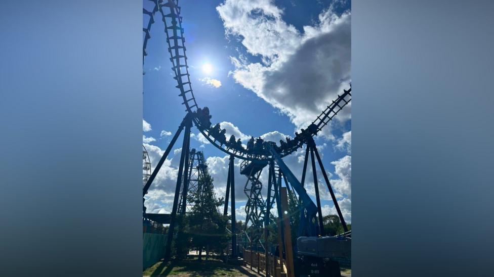 A blue rollercoaster with people suspended at a height in carriages. A blue sky with white clouds is in the background