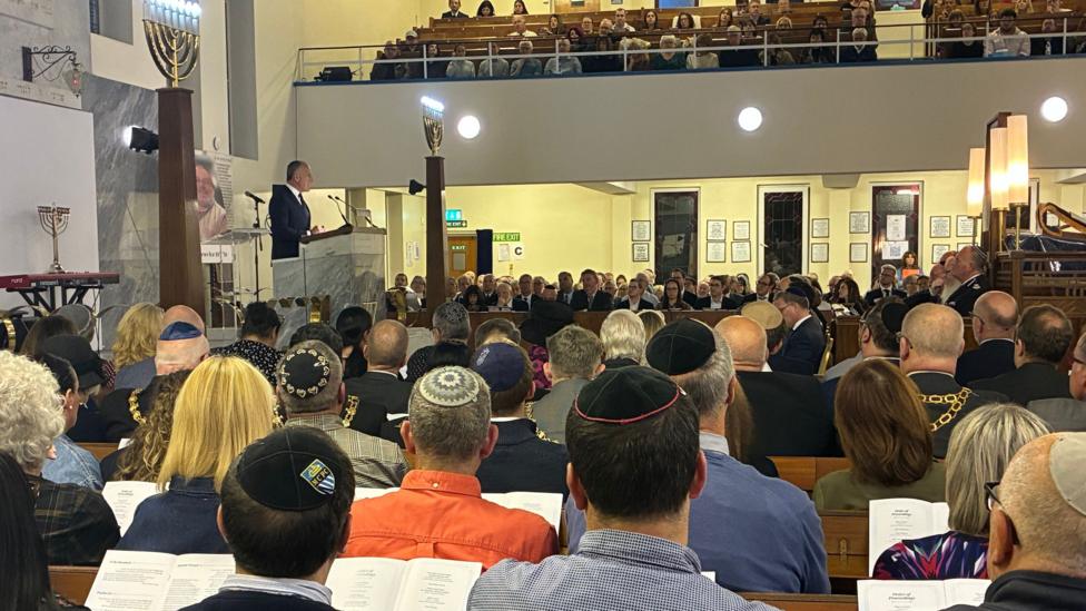Rows of people are seated in the synagogue as Raphi Bloom, wearing a dark suit and tie, addresses them from a high lectern with two microphones. People can also be seen seated in a gallery above.