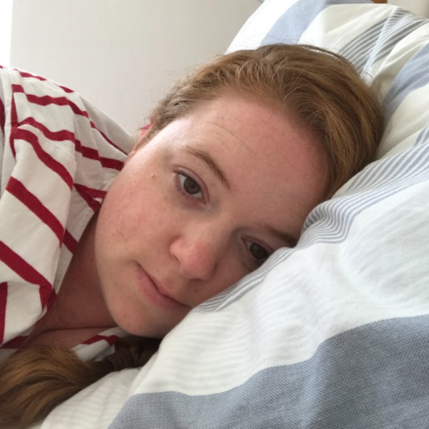 Woman with red hair lying in bed, facing the camera with a sad expression. 