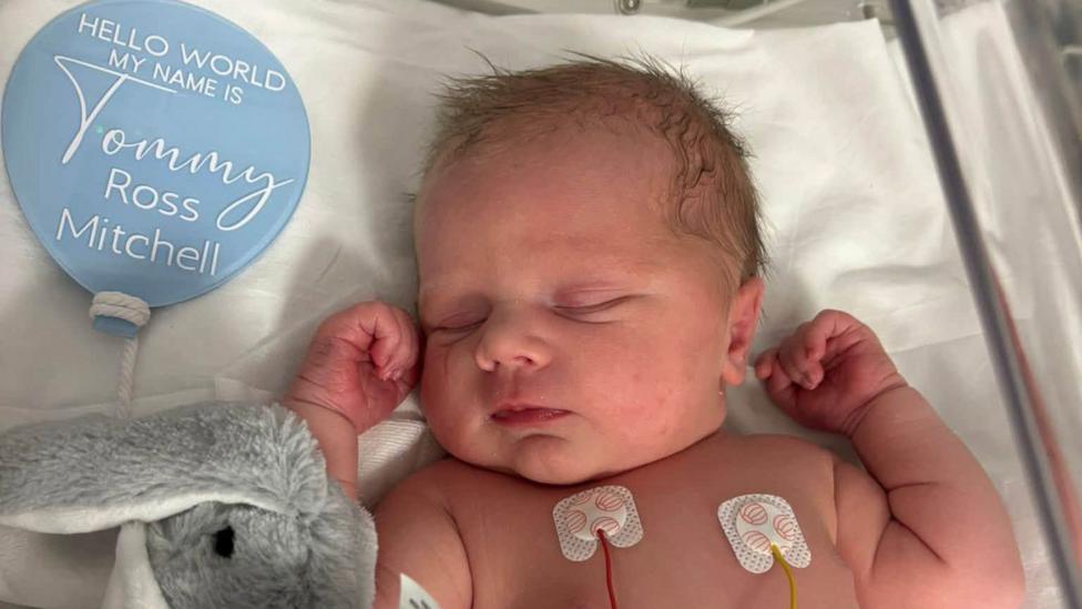 Newborn baby Tommy in a cot. He has his hands up next to his head, his eyes closed and soft toy rabbit next to him. A blue balloon reads his name.