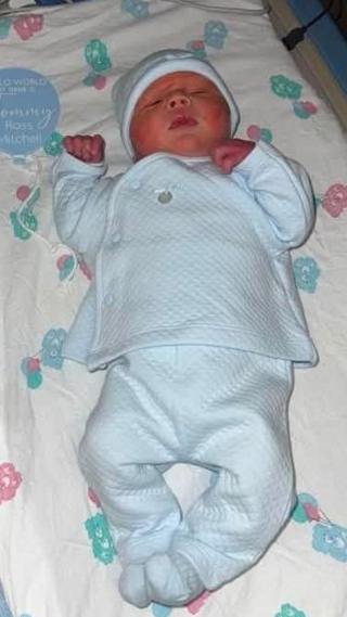 Newborn baby Tommy in a cot. He has his hands up next to his head, his eyes closed and he is wearing a blue sleep suit.