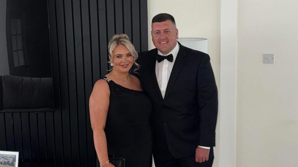Chelsea McGillivray wearing a black dress with her partner Jack who is in a black suit, white shirt and black bow tie. They are standing together, smiling at the camera.