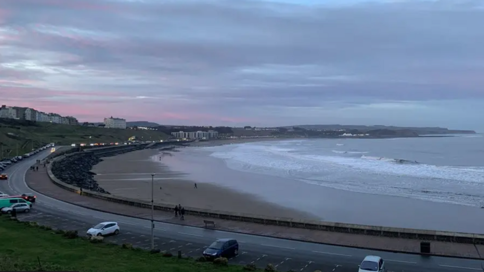 This is an early evening shot of Scarborough's North Bay. The sky is a striking mix of purple and pink. The tide is out and a small number of people are walking on the beach. Traffic is light along North Marine Road and the cars have their headlights on.