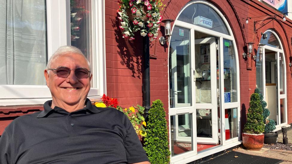 A man sitting in the sunshine outside a B&B in Scarborough. He is smiling at the camera. The sun is on his face and a hanging basket, full of pink and white flowers, is attached to a lamppost behind him.