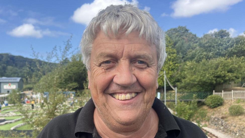 Keith Southwell - a middle-aged man is smiling and looking directly at the camera. Be has blue eyes and thick, short grey hair. You can only see his shoulders and he is wearing a black T-shirt. There is blue sky and trees in the background as well as part of a mini golf course.