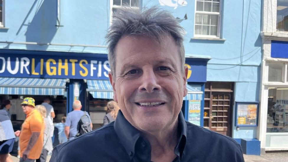 Pete Fraser - a middle-aged man is looking directly at the camera and smiling. He has short grey spiky hair, brown eyes and a slight gap between his two front teeth. He is wearing a navy shirt and standing in front of a blue building that is a fish and chip shop. There are a few people queueing outside in the sun.