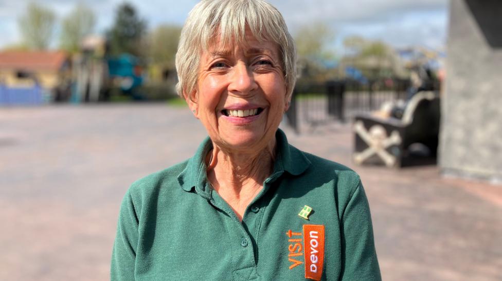 Sally Everton is wearing a green top wit the words Visit Devon on it in orange. She is smiling.
