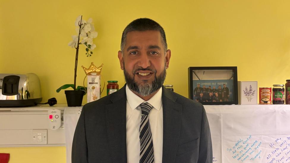 Zameer Ali smiling at the camera, he is standing in the warehouse break room. He is wearing a suit and tie. 