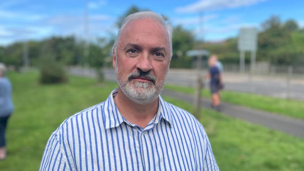 A grey-haired man with grey beard and striped shirt looks at the camera