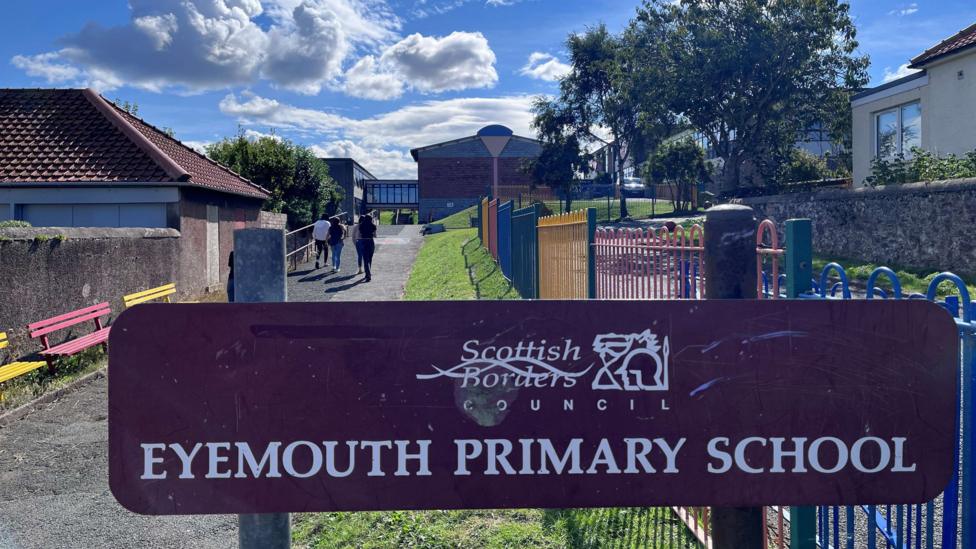 A maroon sign saying Eyemouth Primary School in white lettering with colourful railings and buildings in the background