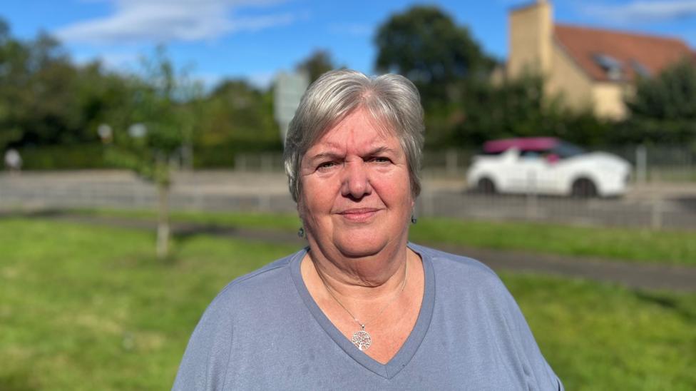 A grey-haired woman wearing a grey top looks at the camera with a grassy area and road in the distance