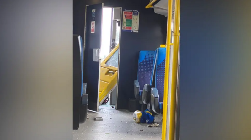 Interior of a passenger train carriage showing debris scattered on the floor and a damaged yellow train cab door hanging open, with blue seats and yellow handrails visible in the foreground.